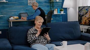 Happy old senior grandmother holding digital tablet browsing, searching informations, shopping online, while adult man working at laptop in background. Person enjoying using notepad gadget photo