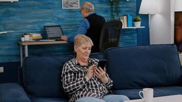 Middle aged woman relaxing holding smartphone reading e book sitting on sofa at home, while senior adult man working at laptop in background. Person enjoying using notepad browsing internet shopping photo