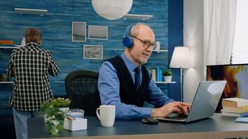 Senior old man listening music on headphones while working on laptop in living room and wife drinking coffee in background. Retired man using modern technology reading typing, searching on computer photo