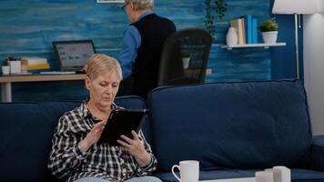 Middle aged old woman relaxing holding tablet reading e book sitting on sofa at home, while senior adult man working at laptop in background. Person enjoying using notepad browsing internet shopping photo