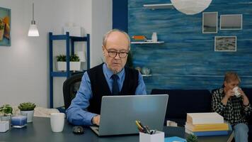 retirado proyecto gerente trabajando desde hogar, apertura ordenador portátil leyendo noticias, mecanografía y analizando financiero Estadísticas, mientras su esposa Bebiendo té en antecedentes. antiguo hombre utilizando moderno tecnología para trabajo foto
