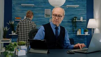 Elderly man using tablet and laptop in same time compering financial graphs working from home sitting in workplace. Busy focused old employee writing, searching, analysing reports photo