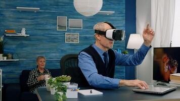 Senior man using virtual reality goggles in living room gesturing sitting in home workplace. Retired man experiencing vr headset while woman watching tv sitting on sofa in background. photo