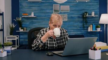 mayor mujer disfrutando un taza de café mientras trabajando en ordenador portátil en vivo habitación mientras marido sentado en el sofá y leyendo libro en antecedentes. maduro más viejo dama acecho en línea negocio formación foto