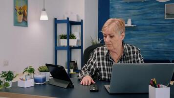 maduro mujer utilizando tableta y ordenador portátil en mismo hora analizando financiero gráficos trabajando desde hogar sentado en lugar de trabajo. ocupado enfocado antiguo empleado escribiendo, buscando, analizando informes foto