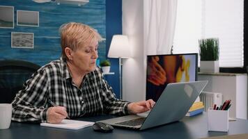 Senior stylish woman taking notes in notebook while using laptop at home. Old freelancer writing details on book while working from workspace in living room, checking financial project of company photo