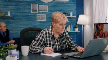 Focused stylish mature old woman remote working from home distance office on laptop taking notes. Middle aged business lady using computer watching webinar sitting on chair writing in notebook. photo