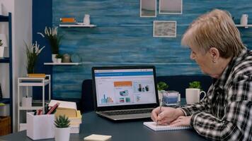 Focused senior entrepreneur checking graphics from laptop writing on notebook working from business office overtime. Busy employee using modern technology network wireless reading typing, searching photo