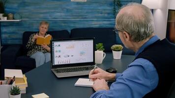 Tired elderly entrepreneur checking graphics from laptop writing on notebook working from business office overtime. Busy employee using modern technology network wireless reading typing, searching photo