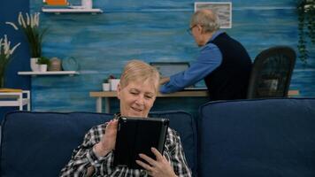 Senior woman reading emails on digital tablet sitting on sofa at home, while senior adult man working at laptop in background. Person enjoying using notepad browsing internet shopping photo