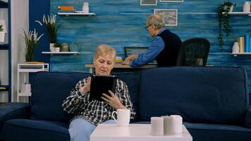 Focused senior grandmother relaxing sitting on sofa typing tablet browsing, searching informations, shopping online, while adult man working at laptop in background. Person using notepad gadget photo