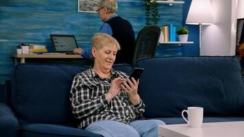 Happy middle aged mature woman enjoying using mobile apps texting typing messages sit on sofa, smiling old adult lady holding smartphone looking at cellphone screen browsing social media at home photo
