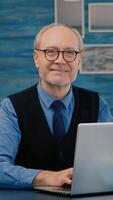 Retired manager sitting at desk in front of camera smiling after typing at laptop working from home while senior wife reading a book in background. Man prepared for online meeting using moden gadgets photo