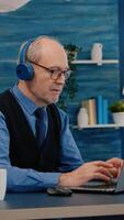 Focused old entrepreneur with headphones listening music while checking graphics writing on laptop working from home. Retired man using modern technology reading typing, searching on computer photo