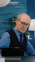 Senior man using laptop and tablet in same time working in office living room at project. Busy focused old employee writing, searching, analysing reports while elderly wife watching tv in background photo