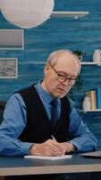 Multitasking retired man reading on laptop and writing on notebook working from home. Focused old remote employee searching, analysing financial reports while elderly wife watching tv in background photo