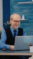 Happy old manager receving good news on laptop working from home for startup company, gesturing victory. Retired employee using modern technology reading typing searching while senior wife watching TV photo