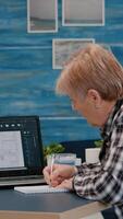 Senior woman architect analysing digital prototype with plans from laptop working from home, taking notes on notebook. Elderly designer using cad software to create a 3D concept of buildings photo