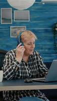Happy old woman entrepreneur with headset listening music while checking statistics, writing on laptop working from home. Retired worker using modern technology reading typing, searching on computer photo