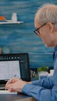 Senior man architect analysing digital prototype with plans from laptop working from home, taking notes on notebook. Elderly engineer designer using cad software to create a 3D concept of buildings. photo