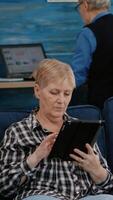 Middle aged old woman relaxing holding tablet reading e book sitting on sofa at home, while senior adult man working at laptop in background. Person enjoying using notepad browsing internet shopping photo