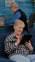 Happy old senior grandmother holding digital tablet browsing, searching informations, shopping online, while adult man working at laptop in background. Person enjoying using notepad gadget photo