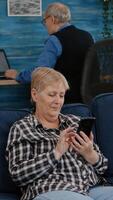 Middle aged woman relaxing holding smartphone reading e book sitting on sofa at home, while senior adult man working at laptop in background. Person enjoying using notepad browsing internet shopping photo