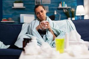 Sick caucasian woman sitting in bed with severe cold and flu drinking hot tea. Adult wrapped in blanket on the sofa with health fever infection symptomps. Person with disease at home, temperature and cough photo