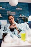 Sick caucasian woman sitting in bed with severe cold and flu drinking hot tea. Adult wrapped in blanket on the sofa with health fever infection symptomps. Person with disease at home, temperature and cough photo
