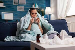 Woman feeling sick in blanket with warm drink at home having seasonal virus symptoms. Unwell young adult in pain headache resting on couch with medical treatment against coronavirus photo