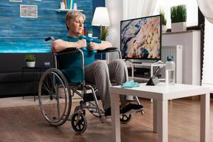 Invalid senior woman in wheelchair training body muscles using gymnastic dumbbells recovery after paralysis. Focused disability pensioner watching gym workout exercise on tablet exercising resistance photo