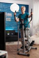 Focused senior woman working muscle legs doing body exercise using cycling bicycle machine during fitness workout. Retirement active pensioner watching aerobics on television in living room photo