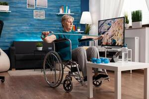 Disabled pensioner in wheelchair working with elastic band training body muscle recovering after disability accident looking at aerobics on tablet. Pensioner doing healthcare arm exercise photo