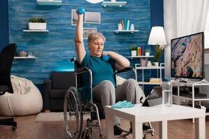 Disabled old woman in wheelchair raising arm training muscles resistance using dumbbells recovery after muscular disability watching cardio on laptop. Pensioner doing gym exercise in living room photo