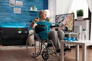 Invalid senior woman in wheelchair holding resistance elastic band stretching body muscle recovering after disability accident watching workout on tablet. Pensioner doing aerobics exercise photo