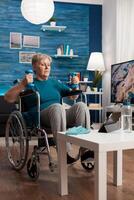Handicapped senior woman holding trainer dumbbells doing arms exercise working at body persistence training in living room. Disabled pensioner looking at online muscles workout course on tablet photo