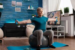 Focused senior woman stretching arm working at body muscle using fitness dumbbells sitting on swiss ball in living room. Caucasian male exercising muscular healthcare during wellness workout photo