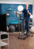 Focused retirement senior man working at legs resistance using cycling bicycle machine watching aerobics on television for well being. Pensioner training body muscle during health cardio workout photo