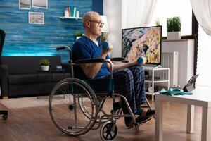 Focused senior man in wheelchair watching online workout on tablet computer in living room working arm muscle using dumbbells. Invalid pensioner recovering body resistance after paralysis photo