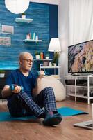 Senior man in sportwear warming up body muscles practicing wellness exercise using workout dumbbells. Pensioner sitting on yoga mat with crossed legs during aerobics routine looking at gym photo