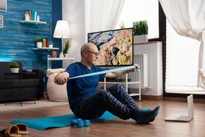 Pensioner working at body resistance exercising arms muscles using elastic band sitting on yoga mat with crossed legs position. Senior man doing workout during fitness class looking on laptop photo