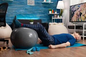 Retirement senior man doing wellness warming legs up using swiss ball sitting on yoga mat in living room. Retired pensioner practicing abdominals exercises working at body muscular healthcare photo