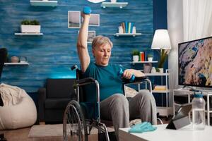 Handicapped senior woman holding trainer dumbbells doing arms exercise working at body persistence training in living room. Disabled pensioner looking at online muscles workout course on tablet photo