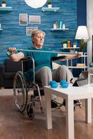 Invalid senior woman in wheelchair holding resistance elastic band stretching body muscle recovering after disability accident watching workout on tablet. Pensioner doing aerobics exercise photo