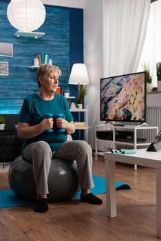 enfocado mayor hombre hacer ejercicio cuerpo resistencia rutina de ejercicio haciendo brazos ejercicio utilizando pesa sentado en aptitud suizo pelota en vivo habitación. pensionista acecho en línea bienestar formación en ordenador portátil foto