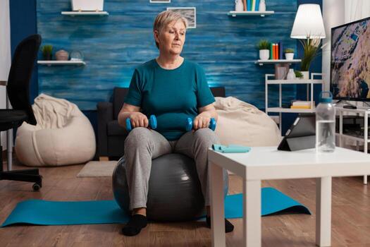 mayor mujer hacer ejercicio cuerpo músculos haciendo brazos ejercicio sentado en suizo pelota en vivo habitación. pensionista acecho en línea aeróbicos formación en tableta hacer ejercicio muscular resistencia participación pesa foto