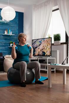 enfocado mayor hombre participación pesas trabajando músculos ejercicio estirar cuerpo formación brazo muscular resistencia. Jubilación caucásico masculino sentado en suizo pelota en vivo habitación haciendo aptitud rutina de ejercicio foto