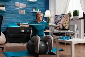 Retired senior man sitting on fitness swiss ball in living room doing wellness fitness workout streching arm muscles using aerobics elastic band. Pensioner training body resistance in living room photo