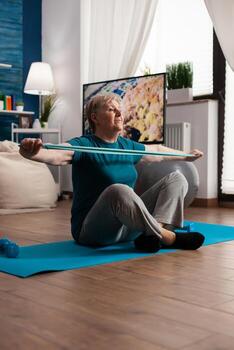 Jubilación mayor mujer sentado en yoga estera extensión piernas músculos utilizando tramo elástico banda formación cuerpo resistencia. pensionista en ropa de deporte adelgazar peso durante músculo formación en vivo habitación foto