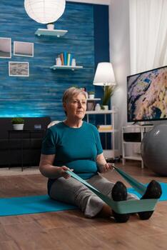 enfocado pensionista vistiendo ropa de deporte sentado en yoga estera extensión piernas músculo utilizando aptitud elástico banda durante aeróbicos rutina de ejercicio hacer ejercicio músculos resistencia. mayor mujer haciendo cuerpo ejercicio foto